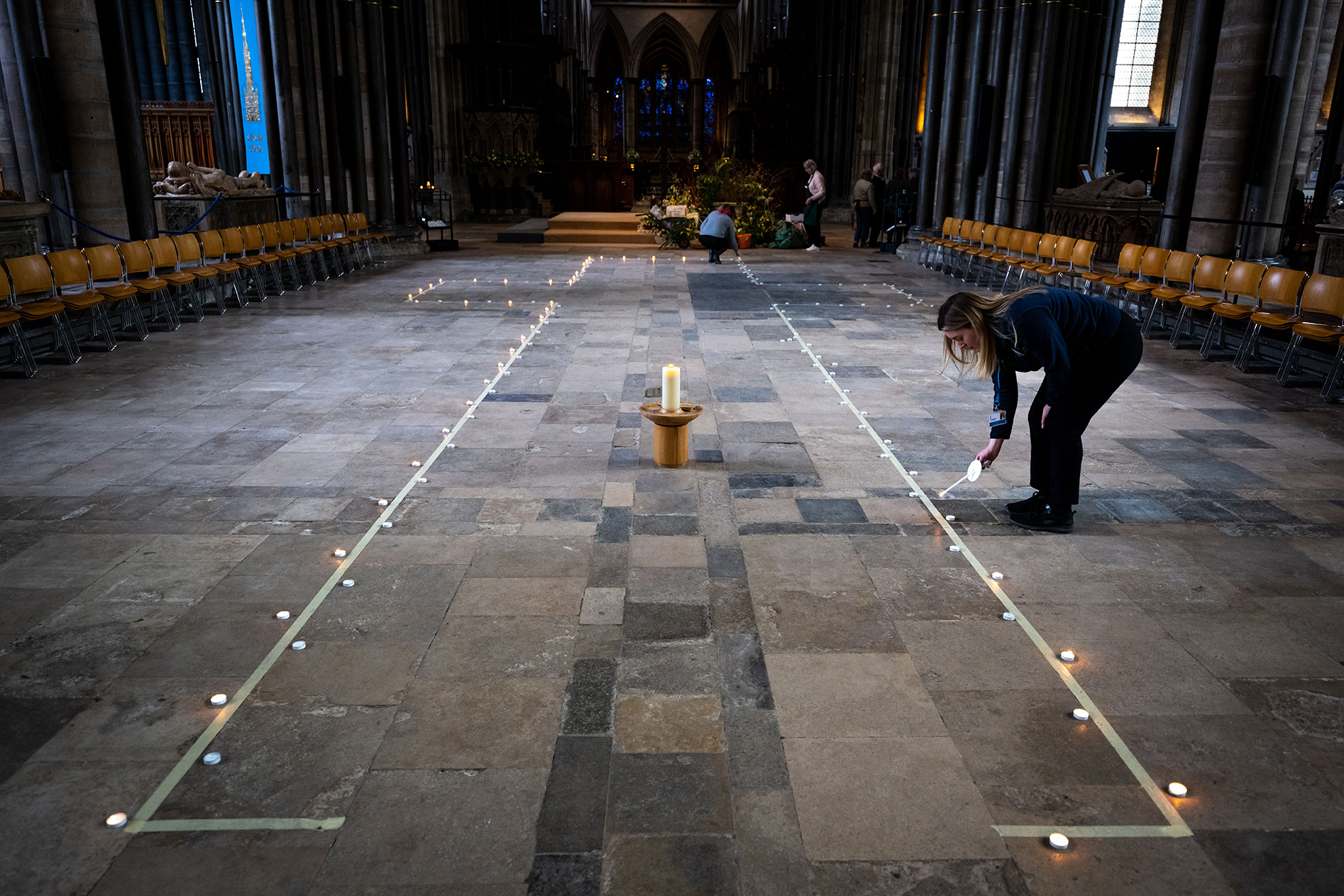 A line of candles marking out a cross on the floor. A woman is lighting a candle on the edge.